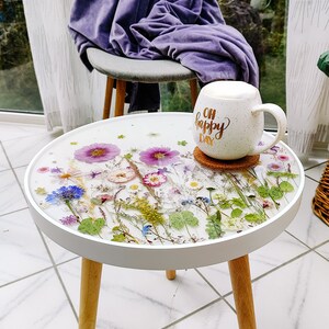 Puede incluir: Mesa auxiliar redonda blanca con una tapa blanca y patas de madera. La tapa est&aacute; decorada con flores y vegetaci&oacute;n prensadas en una resina transparente. Una taza blanca con el texto "Oh Happy Day" est&aacute; encima de la mesa.