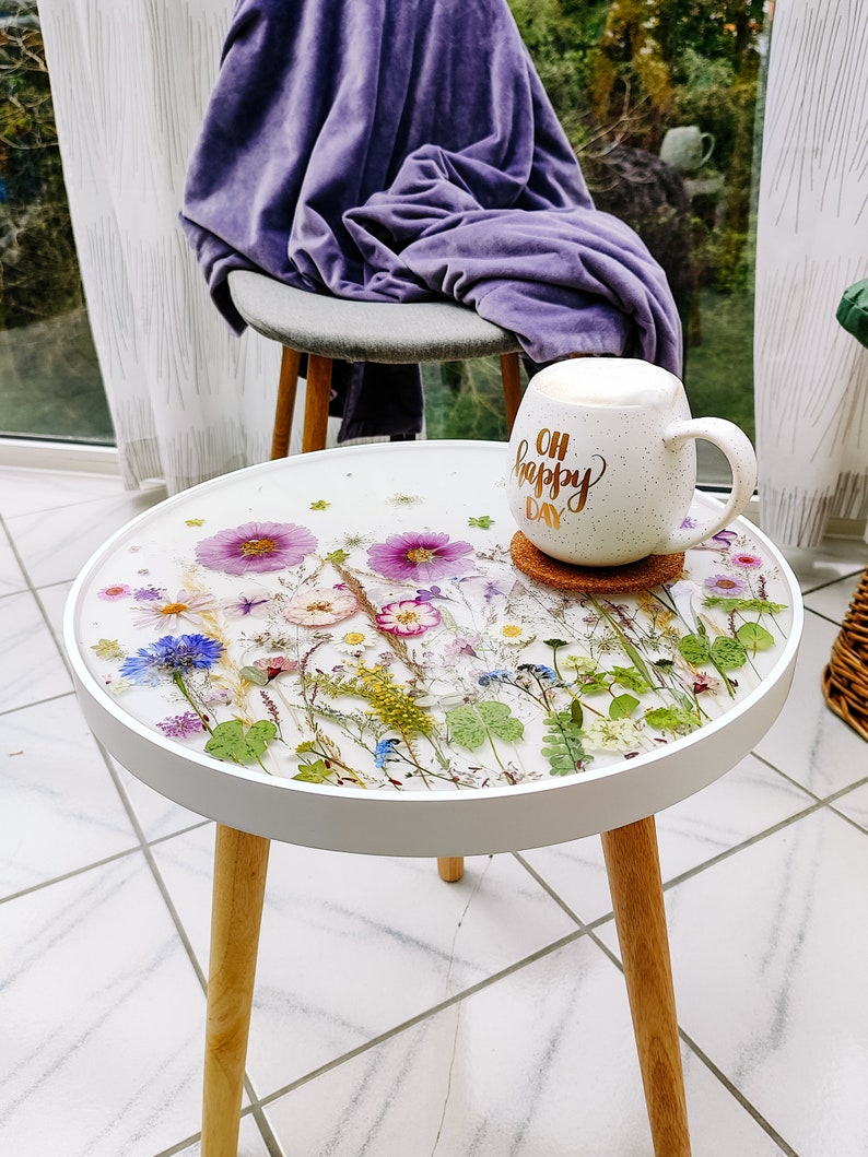 Puede incluir: Mesa auxiliar redonda blanca con una tapa blanca y patas de madera. La tapa est&aacute; decorada con flores y vegetaci&oacute;n prensadas en una resina transparente. Una taza blanca con el texto "Oh Happy Day" est&aacute; encima de la mesa.