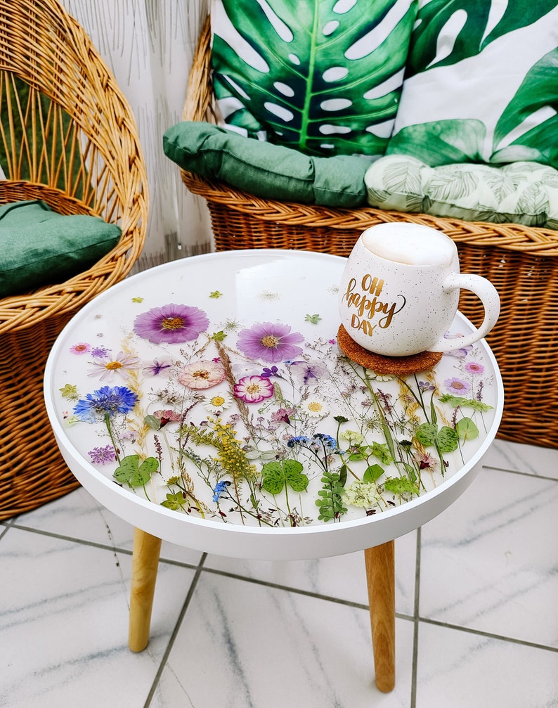 Puede incluir: Mesa auxiliar redonda blanca con base de madera. La superficie de la mesa est&aacute; decorada con flores y vegetaci&oacute;n prensadas. Una taza blanca con el texto "Oh Happy Day" est&aacute; sobre la mesa.