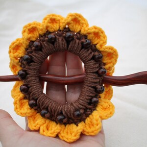 Puede incluir: Un palo de pelo de madera marr&oacute;n con un dise&ntilde;o de girasol tejido a crochet. El girasol est&aacute; hecho con hilo marr&oacute;n y amarillo y tiene cuentas de madera marr&oacute;n alrededor del centro.