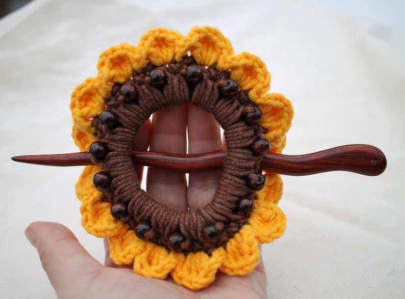 Puede incluir: Un palo de pelo de madera marr&oacute;n con un dise&ntilde;o de girasol tejido a crochet. El girasol est&aacute; hecho con hilo marr&oacute;n y amarillo y tiene cuentas de madera marr&oacute;n alrededor del centro.