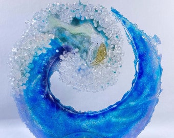 Puede incluir: Una escultura de vidrio azul y blanco de una ola con una figura en su interior. La escultura está sobre un soporte de vidrio transparente.