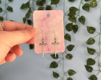 May include: Silver-colored earrings with a sword design. The earrings are displayed on a pink and white card. The background is a light blue wall with green vines.