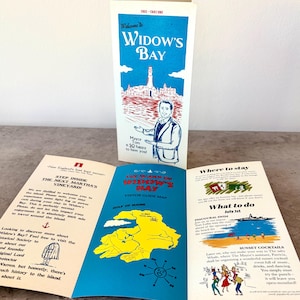 May include: A tri-fold visitor guide brochure for Widow's Bay featuring a blue and red graphic of a lighthouse and a mayor on the cover. The interior displays a yellow map of the island, text about local events, and a guide to staying.