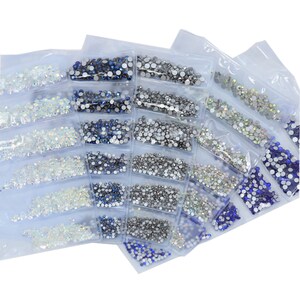 May include: Assortment of clear, silver, and blue rhinestones in individual plastic bags. The rhinestones are small and round, and they are arranged in rows in the bags.