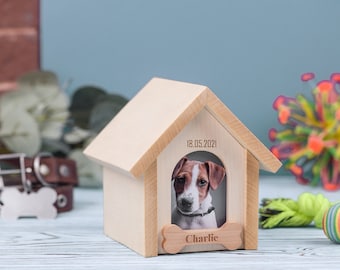 Puede incluir: Una casa de perro de madera con una foto de un perro marrón y blanco en el interior. La casa de perro tiene una placa en forma de hueso con el nombre "Charlie" y la fecha "18.05.2021" grabada en ella.