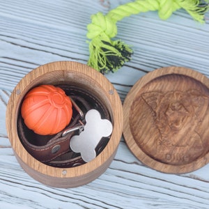 May include: A wooden box with a carved dog on the lid. The box is open and contains a leather dog collar, a bone-shaped dog tag, and an orange rubber ball.