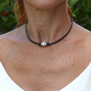 May include: A dark brown leather choker necklace with a single, round, white pearl pendant. The pearl is centered on the necklace and secured with silver-colored metal accents. The necklace is worn around the neck.