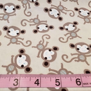May include: Tan fabric with a repeating pattern of cartoon monkeys in various poses. The monkeys are brown with white faces, blue accents, and dark brown ears. A pink measuring tape is across the bottom of the fabric.
