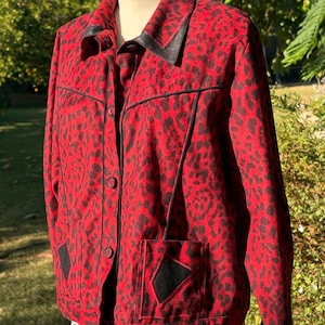 Op de afbeelding: Een rood en zwart luipaardprint knoopjasje met zwarte bies en een zwarte ruitvormige patch op de zak.