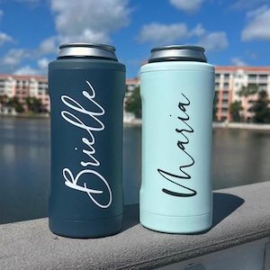 May include: Two personalized can coolers. One is a dark teal color with the name "Brielle" in white script. The other is a light blue color with the name "Maria" in white script. Both coolers have a silver rim and are cylindrical in shape.