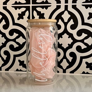 Peut inclure: Un bocal en verre transparent avec un couvercle en bambou, rempli de papier de soie rose. Le mot "Mallory" est &eacute;crit en blanc sur le devant du bocal. Le bocal est pos&eacute; sur une surface blanche devant un fond de carrelage &agrave; motifs noir et blanc.