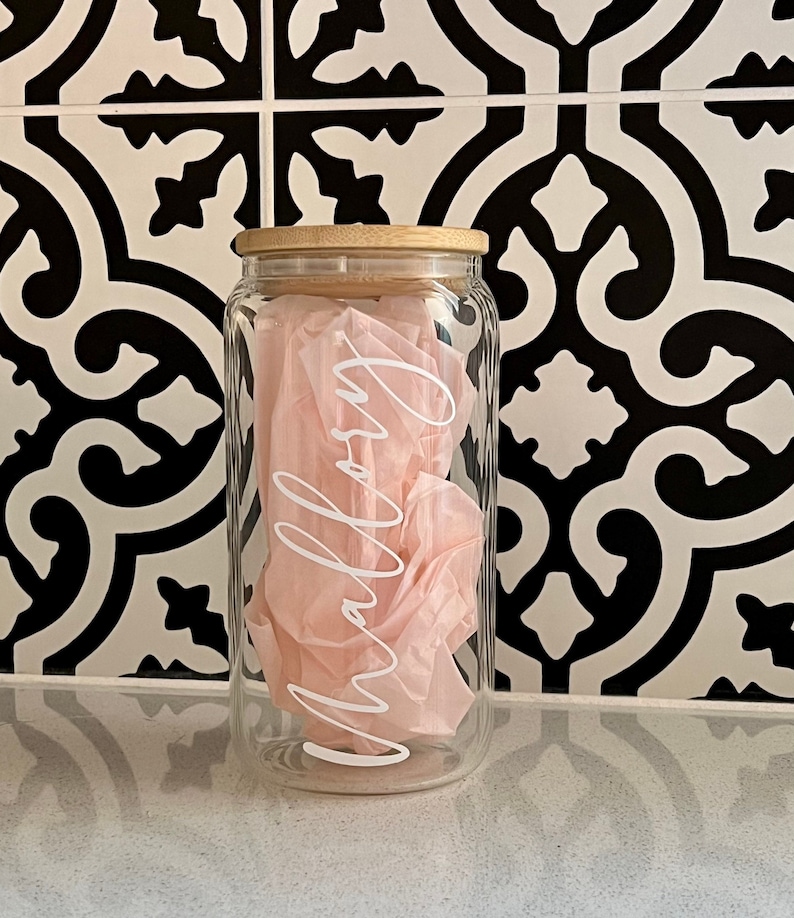 Peut inclure: Un bocal en verre transparent avec un couvercle en bambou, rempli de papier de soie rose. Le mot "Mallory" est &eacute;crit en blanc sur le devant du bocal. Le bocal est pos&eacute; sur une surface blanche devant un fond de carrelage &agrave; motifs noir et blanc.