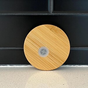 Peut inclure: Couvercle rond en bambou brun clair avec un petit bouchon en plastique transparent au centre. Le couvercle pr&eacute;sente un motif de grain de bois naturel et est con&ccedil;u pour un verre ou une bouteille. Le fond est un carrelage noir.
