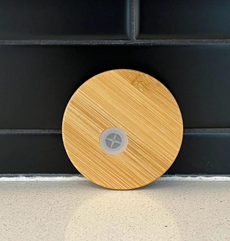 Peut inclure: Couvercle rond en bambou brun clair avec un petit bouchon en plastique transparent au centre. Le couvercle pr&eacute;sente un motif de grain de bois naturel et est con&ccedil;u pour un verre ou une bouteille. Le fond est un carrelage noir.