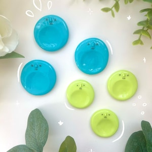 May include: A selection of ceramic dishes in turquoise and lime green, each with a minimalist face design. The dishes are round with a shallow bowl shape. The background is white with decorative elements and greenery.