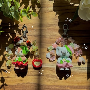 May include: Two keychains featuring flocked animal figures. The left keychain has a deer in a red strawberry-patterned outfit with a red apple charm. The right keychain has an elephant in a pink outfit with translucent flower and butterfly beads.