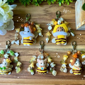 May include: Five keychains featuring small flocked animal figures dressed in yellow and black striped bee costumes. Each figure is adorned with various beads, including flower-shaped, translucent, and pearl-like accents, attached to a silver-toned metal lobster clasp.