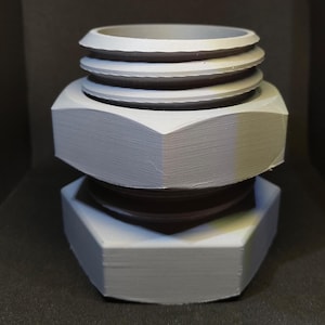 Op de afbeelding: Een wit 3D-geprint plastic model van een grote moer met een kleinere moer erop. De bovenste moer heeft een cilindrische vorm met drie ribben.