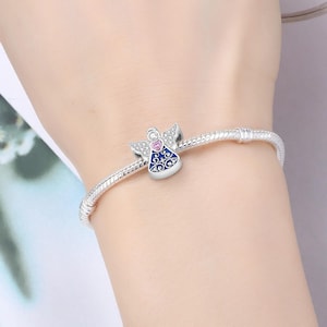 May include: A silver charm bracelet with a detailed angel charm. The angel charm features a pink heart, blue enamel, and clear crystals. The bracelet is worn on a wrist.