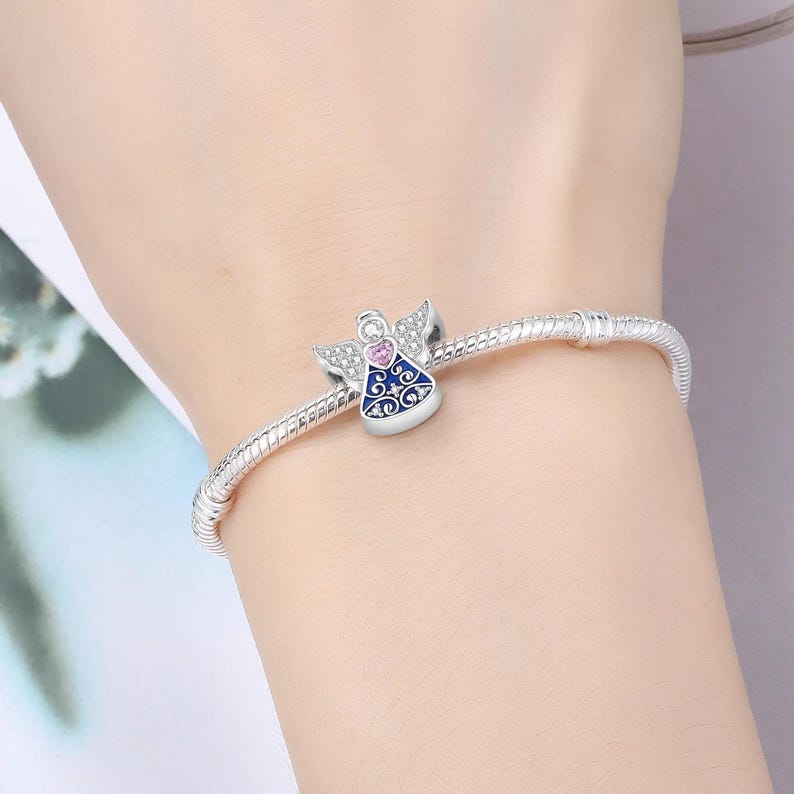 May include: A silver charm bracelet with a detailed angel charm. The angel charm features a pink heart, blue enamel, and clear crystals. The bracelet is worn on a wrist.