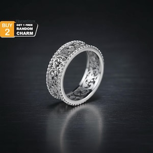 Puede incluir: Un anillo de plata con un intrincado diseño floral. El anillo presenta un diseño detallado alrededor de la banda y una superficie texturizada. El anillo se muestra sobre un fondo oscuro.