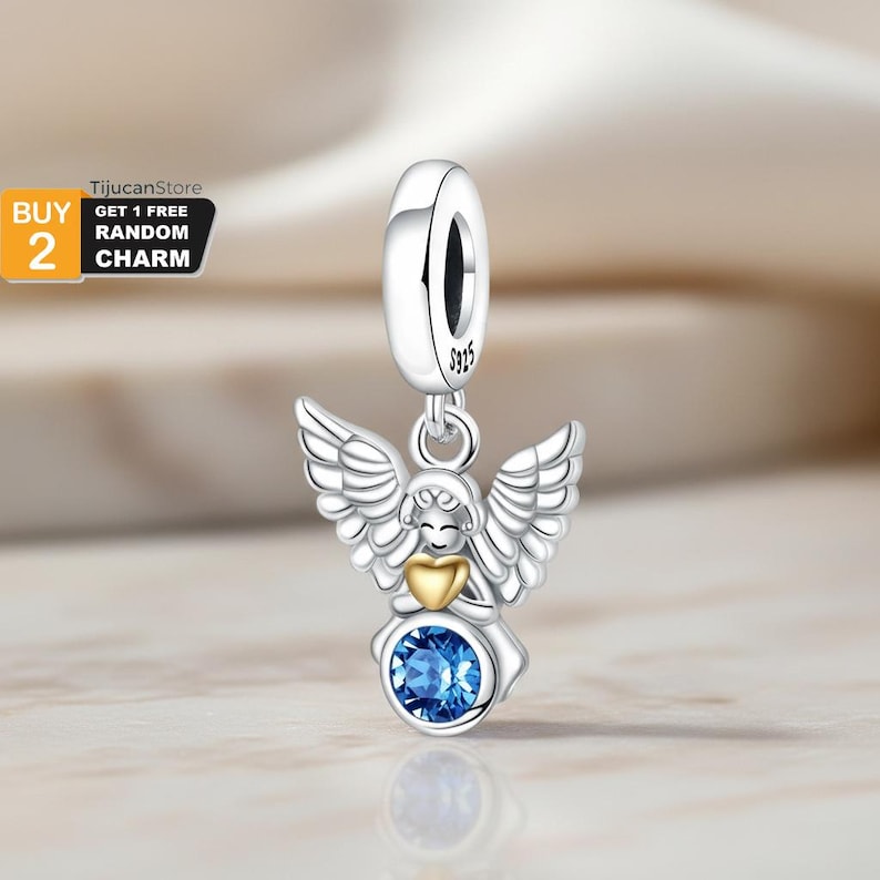 May include: A silver charm with an angel design. The angel has outstretched wings, a gold heart, and a blue gemstone. The charm is suspended from a silver loop. The text "BUY 2 GET 1 FREE RANDOM CHARM" is in the upper left corner.