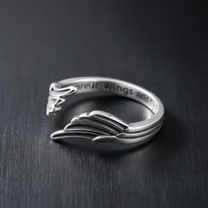 Puede incluir: Un anillo plateado con un dise&ntilde;o abierto, con detalles en forma de alas en cada extremo. El anillo tiene la inscripci&oacute;n "spread your wings and fly" grabada en la banda. El anillo est&aacute; sobre un fondo oscuro.