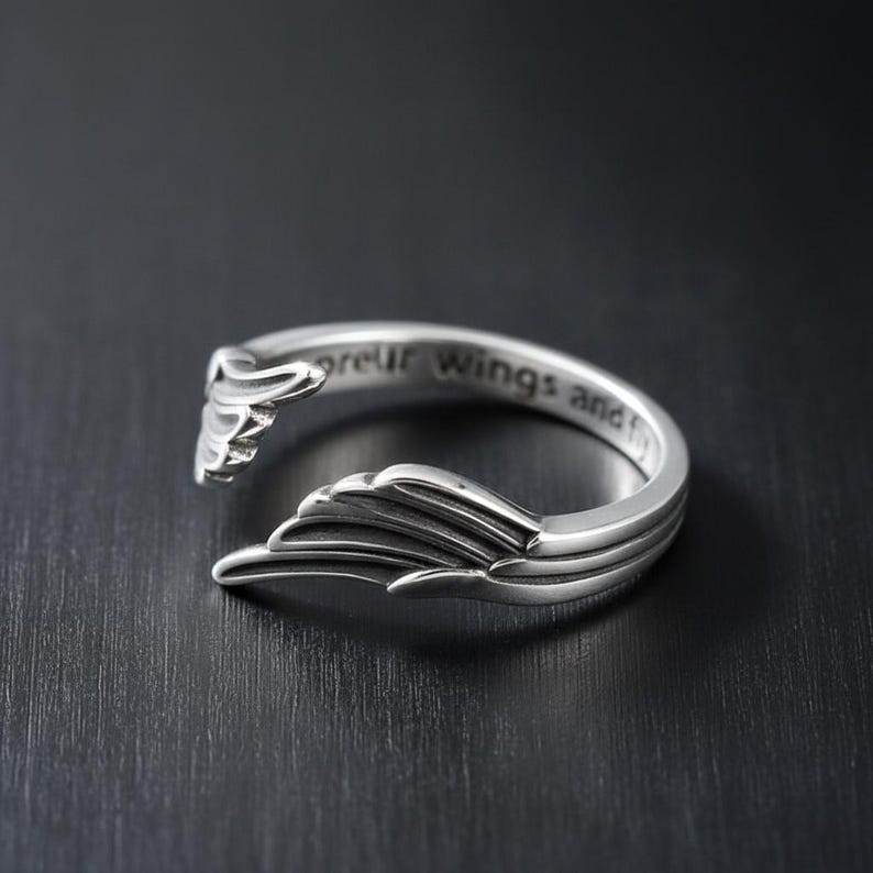 Puede incluir: Un anillo plateado con un dise&ntilde;o abierto, con detalles en forma de alas en cada extremo. El anillo tiene la inscripci&oacute;n "spread your wings and fly" grabada en la banda. El anillo est&aacute; sobre un fondo oscuro.