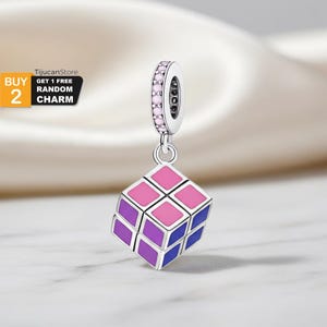 May include: A silver charm with a Rubik's Cube design. The cube features pink, purple, and blue squares. The charm has a ring adorned with small pink stones and the text "BUY 2 GET 1 FREE RANDOM CHARM".