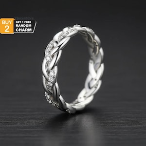 May include: A silver ring featuring a braided design, embellished with small, sparkling stones. The ring is displayed against a dark backdrop. The image also includes text that says "BUY 2" and "GET 1 FREE RANDOM CHARM".