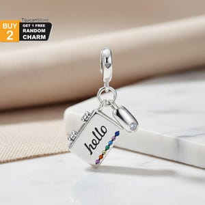 Puede incluir: Un charm plateado con un diseño de cuaderno, la palabra "hello" en escritura negra y una fila de gemas de colores arcoíris. Un pequeño charm plateado con forma de bolígrafo con una gema transparente cuelga junto al cuaderno.