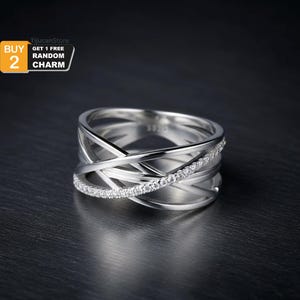 May include: A silver ring with an interwoven design. One band is encrusted with small, clear stones. The ring has a modern, geometric style. The image includes text that says "BUY 2 GET 1 FREE RANDOM CHARM".