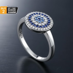 Op de afbeelding: Een zilveren ring met een cirkelvormig ontwerp met een patroon van blauwe en witte stenen. De ring heeft een gladde, gepolijste band en het ontwerp doet denken aan een oog. De ring staat tegen een donkere achtergrond.