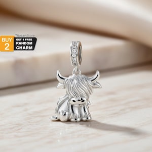 May include: A silver charm in the shape of a Highland cow. The charm has a detailed design with horns, shaggy fur, and a small face. The charm is suspended from a ring adorned with small, clear stones. The text "BUY 2 GET 1 FREE RANDOM CHARM" is in the upper left corner.