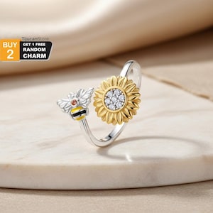 May include: A silver ring featuring a sunflower and a bee design. The sunflower has a gold-colored center with clear stones and yellow petals. The bee has silver wings, a red stone, and a yellow and black striped body. The ring is on a white surface.