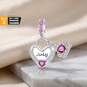 Può includere: Un charm in argento con un ciondolo a forma di cuore con la parola July scritta in nero, accentuato da una piccola pietra rosa a forma di cuore. Un charm a forma di corona con una pietra rosa pende accanto e la maglia è rosa.