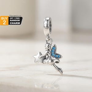 Puede incluir: Un charm plateado con una hada sosteniendo una estrella, con un ala enjoyada azul. El charm tiene una cuenta en forma de estrella en la parte superior. El texto "BUY 2 GET 1 FREE RANDOM CHARM" está en la esquina superior izquierda.