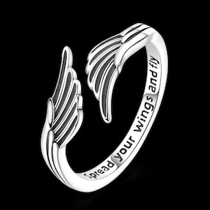 Puede incluir: Anillo plateado con dise&ntilde;o de alas en ambos extremos. El anillo tiene grabadas las palabras "Spread your wings and fly". El anillo est&aacute; sobre un fondo negro. Una joya.
