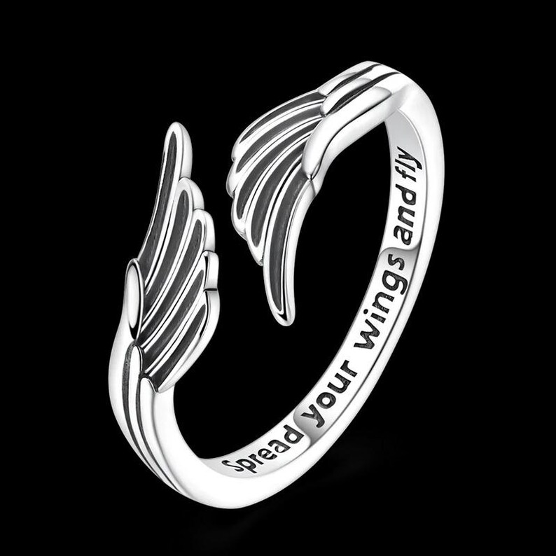 Puede incluir: Anillo plateado con dise&ntilde;o de alas en ambos extremos. El anillo tiene grabadas las palabras "Spread your wings and fly". El anillo est&aacute; sobre un fondo negro. Una joya.