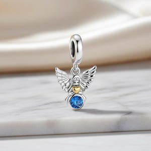May include: A silver angel charm with detailed wings, a gold heart, and a large blue gemstone. The charm is designed to be added to a bracelet or necklace. The angel is suspended from a silver loop.