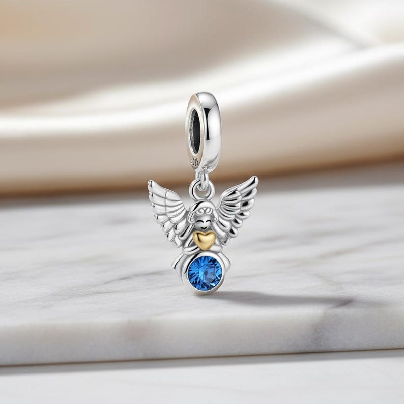 May include: A silver angel charm with detailed wings, a gold heart, and a large blue gemstone. The charm is designed to be added to a bracelet or necklace. The angel is suspended from a silver loop.