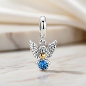May include: A silver angel charm with detailed wings, a gold heart, and a blue gemstone. The charm has a loop at the top for attaching to a bracelet or necklace. The angel is smiling, and the charm is set against a blurred background.