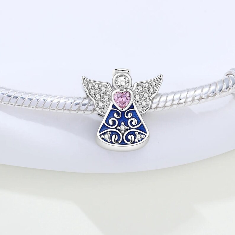 May include: A silver angel charm with a blue enamel dress and a pink heart. The angel has crystal-like wings and a halo. The charm is designed to be added to a bracelet.