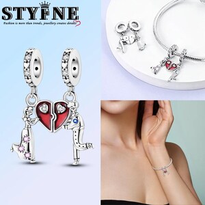 Puede incluir: Pulseras de plata con colgantes en forma de corazón. Un colgante presenta una pareja sosteniendo un corazón rojo, mientras que otro muestra una pareja con un corazón blanco. Los abalorios están adornados con cristales transparentes. Incluye el texto "STYFNE" y "Fashion is more than trends, jewellery creates details."
