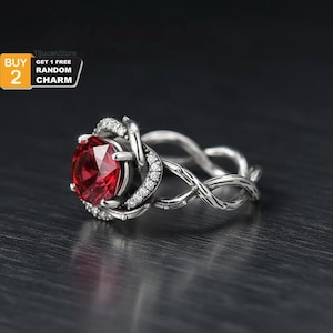 May include: A silver ring with a large, oval-cut red gemstone at the center, surrounded by smaller clear stones. The band features an intricate, interwoven design. The image includes text that says "BUY 2 GET 1 FREE RANDOM CHARM."