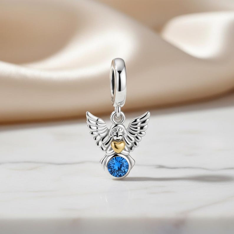 May include: A silver angel charm with detailed wings, a gold heart, and a large, round blue gemstone. The charm has a loop at the top for easy attachment to a bracelet or necklace. The angel charm is a symbol of protection and love.
