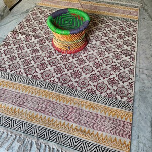 May include: A white cotton rug with a black and brown geometric pattern and fringe. A woven stool with green, blue, red, and yellow stripes sits on the rug.