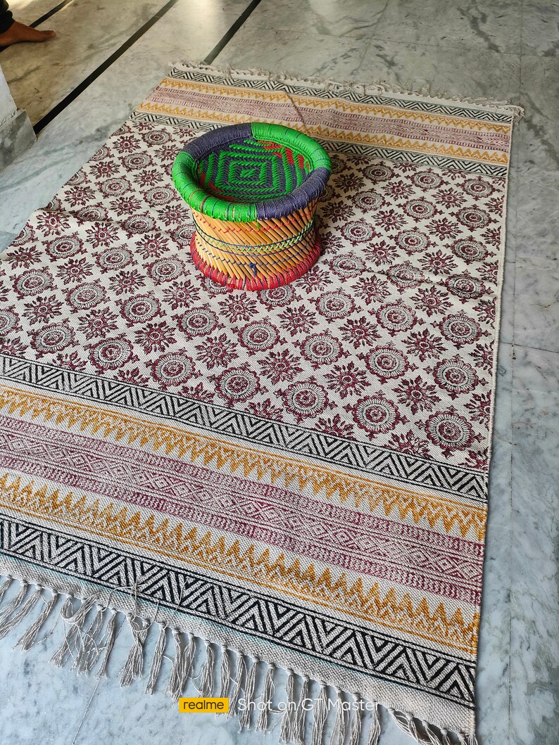 May include: A white cotton rug with a black and brown geometric pattern and fringe. A woven stool with green, blue, red, and yellow stripes sits on the rug.