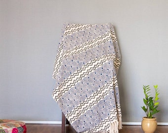 May include: A cream-colored throw blanket draped over a wooden chair. The blanket features a repeating pattern of navy blue half-circles and zigzags. The blanket has a fringe along the bottom edge. A potted plant sits to the right.
